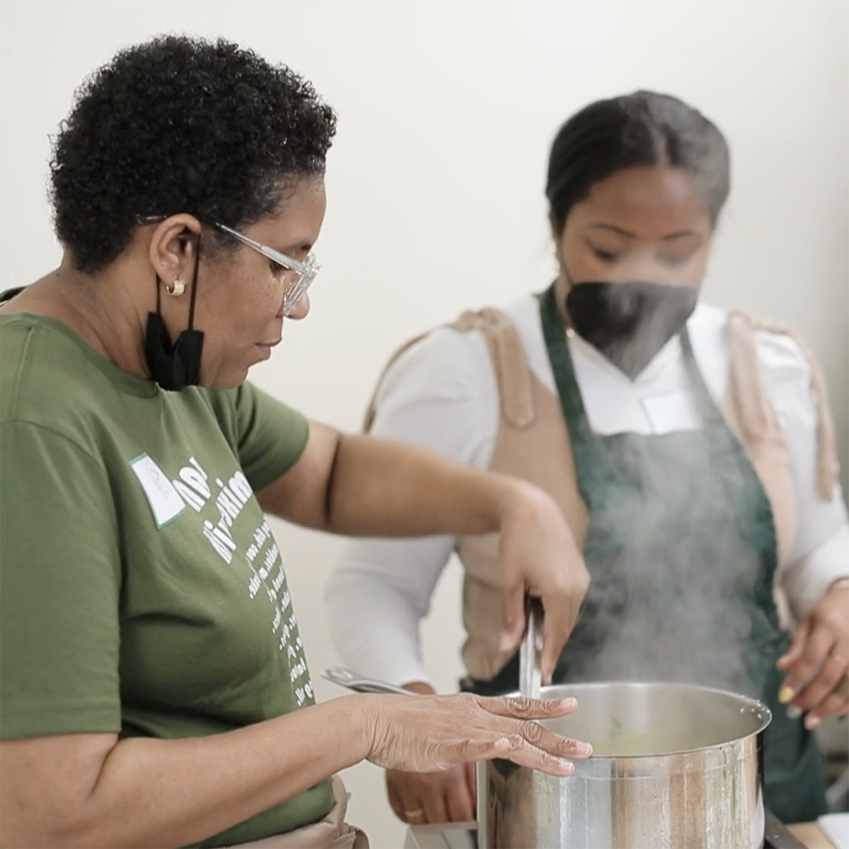 一个黑人妇女的照片, 托尼·辛普森, 在锅里搅拌食物，并向一位年轻的黑人妇女示范，她围着围裙，戴着面具，正在观察