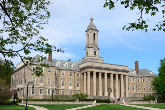 Photo of "Old Main," a grey stone academic hall with a bell tower, at Penn State framed by tree branches with green leaves. 