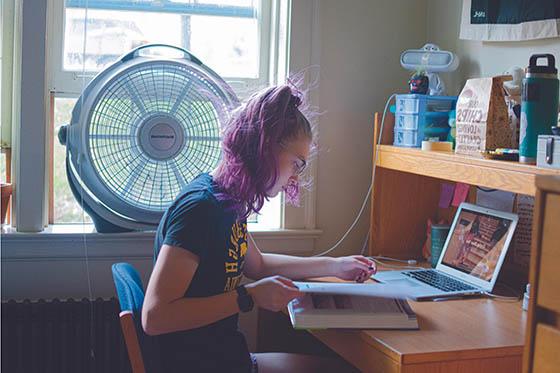A student sits at a desk in her dorm room working at a computer and looking at a text book