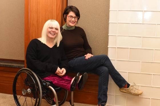 Photo of a woman in a wheelchair and a woman sitting on the arm of it, smiling for the camera