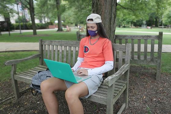 Photo of a Chatham University student in a mask, outside, sitting on a bench and working on her compuer.
