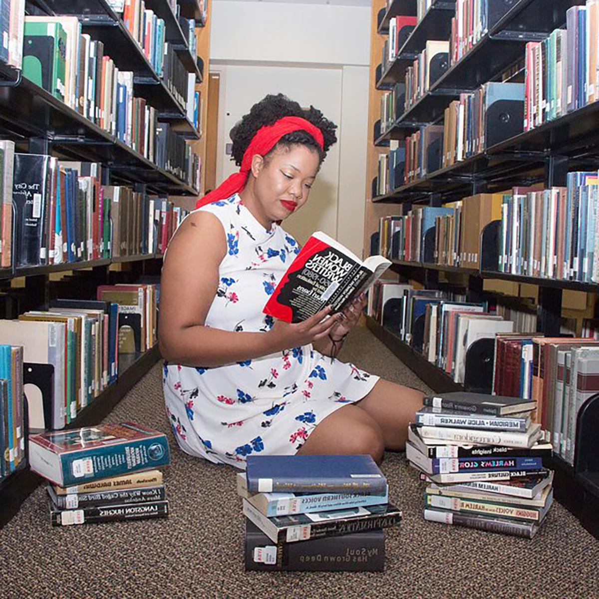 Photo of Caitlyn Hunter, a young Black woman, seated between two library bookshelves with a pile of books in front of her. She is reading a book titled "Black Women Writers, 1950-1980"